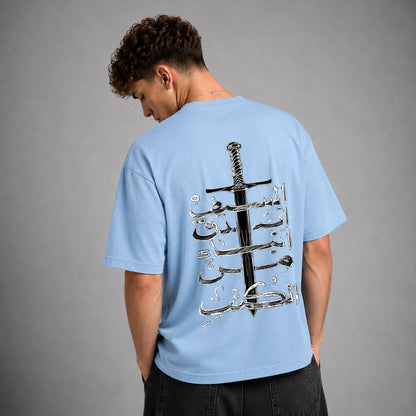A man wearing a light blue t-shirt from the back, featuring a large graphic print of a black sword and silver Arabic calligraphy. The background is a solid gray studio wall.