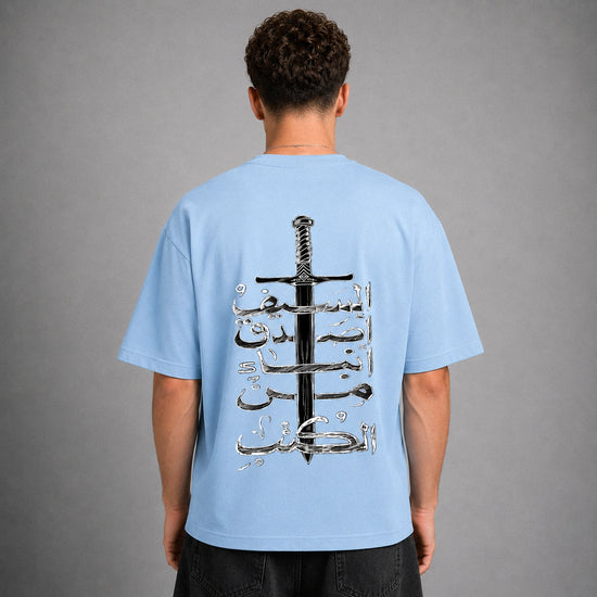 A man viewed from behind wearing a light blue t-shirt with a large graphic print on the back featuring a black sword and silver Arabic calligraphy, set against a plain gray studio background.
