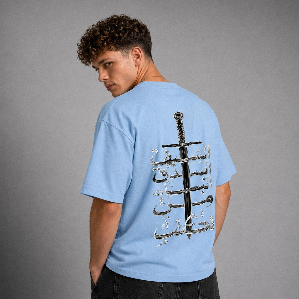 A man wearing a light blue t-shirt from the back, featuring a large graphic print of a black sword and silver Arabic calligraphy on the back, set against a plain gray studio background.