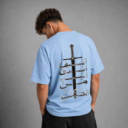 A man wearing a light blue t-shirt from the back, featuring a large graphic print of a black sword and silver Arabic calligraphy on the back, set against a plain gray studio background.