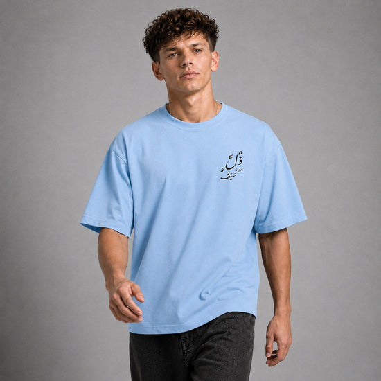 A man wearing a light blue crewneck t-shirt with black Arabic calligraphy on the left chest, paired with dark gray jeans, set against a solid gray studio background.