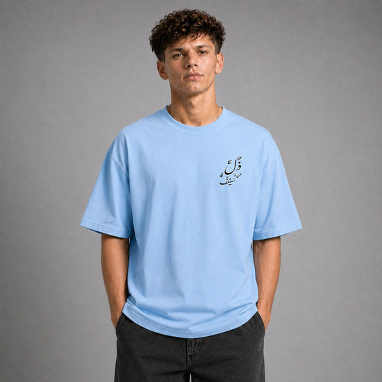 A young man wearing a light blue crewneck t-shirt with a small black Arabic calligraphy graphic on the left chest, paired with dark gray jeans, standing against a neutral gray studio background.