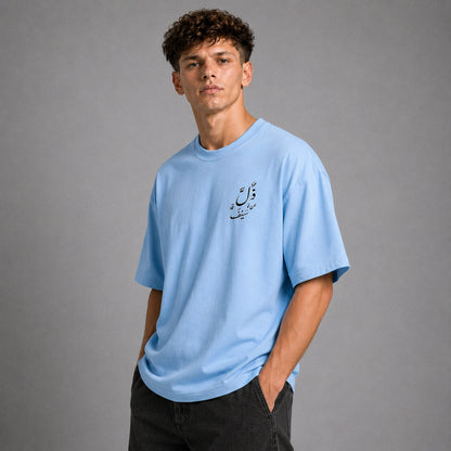 A young man with curly brown hair wearing a light blue crewneck t-shirt with black Arabic calligraphy on the left chest, paired with dark gray jeans, standing against a solid gray studio background.