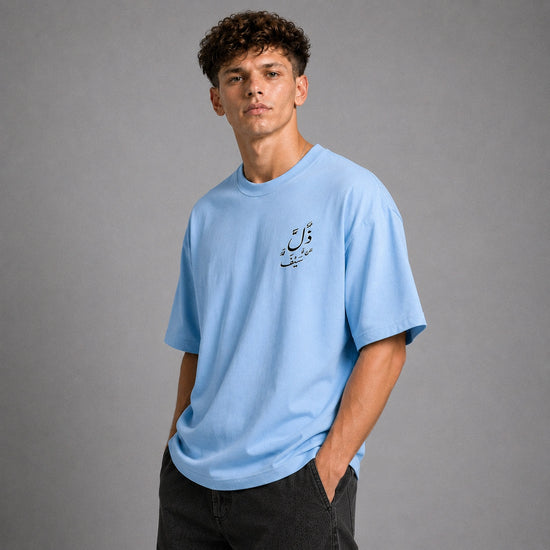 A young man with curly brown hair wearing a light blue crewneck t-shirt with black Arabic calligraphy on the left chest, paired with dark gray jeans, standing against a solid gray studio background.