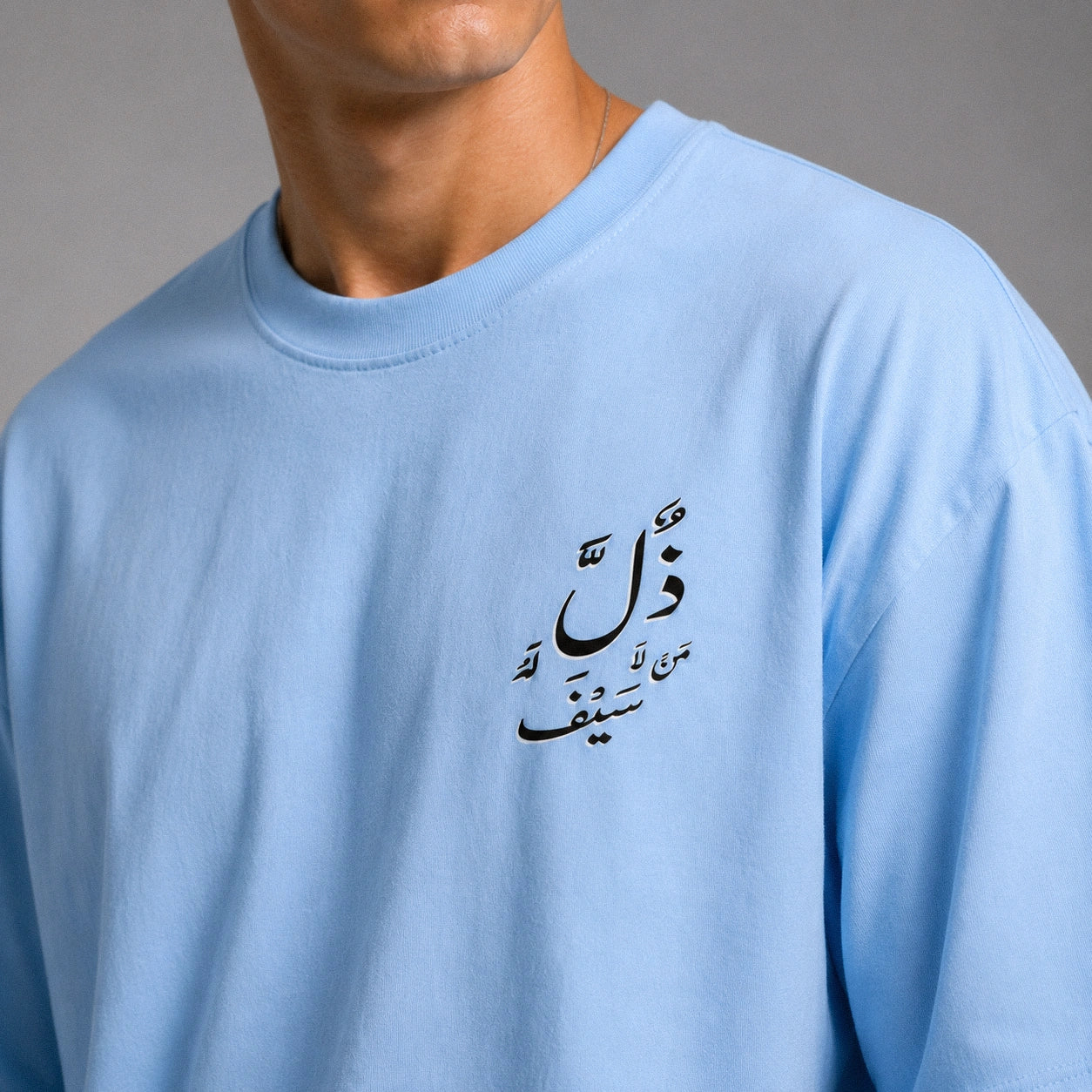 A close-up of a person wearing a light blue crewneck t-shirt with black Arabic calligraphy printed on the left chest, set against a neutral gray background.