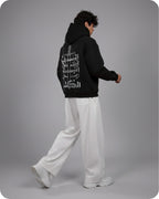 Person wearing a black hoodie with white text on the back, standing against a gray background