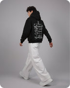 Person wearing a black hoodie with white text on the back, standing against a gray background