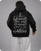 Person wearing a black hoodie with white Arabic calligraphy and sword design on the back, standing against a gray background.