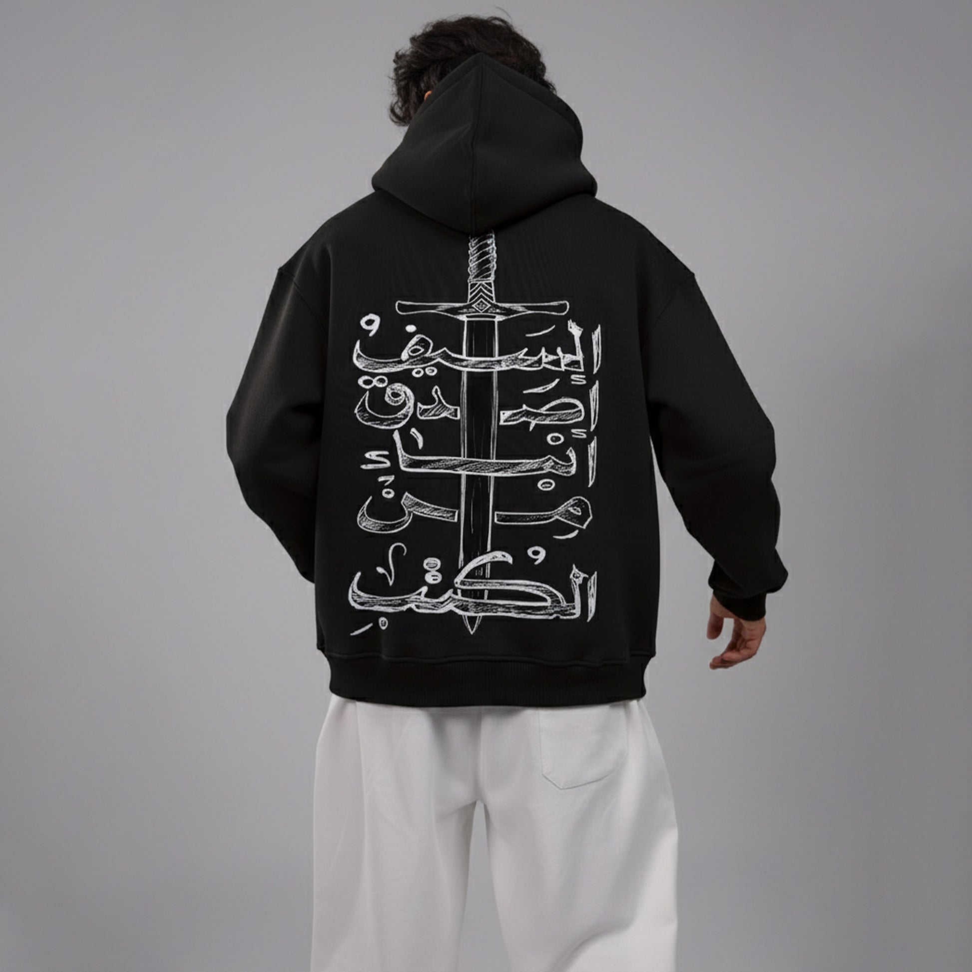 Person wearing a black hoodie with white Arabic calligraphy and sword design on the back, standing against a gray background.