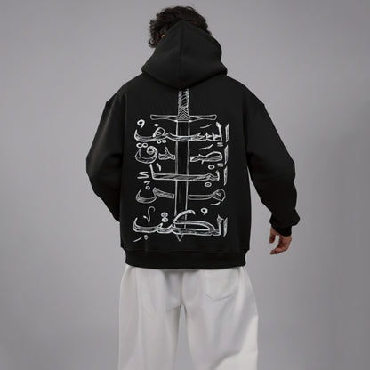 Person wearing a black hoodie with white Arabic calligraphy and sword design on the back, standing against a gray background.