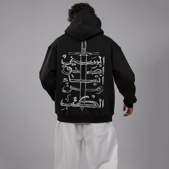 Person wearing a black hoodie with white Arabic calligraphy and sword design on the back, standing against a gray background.