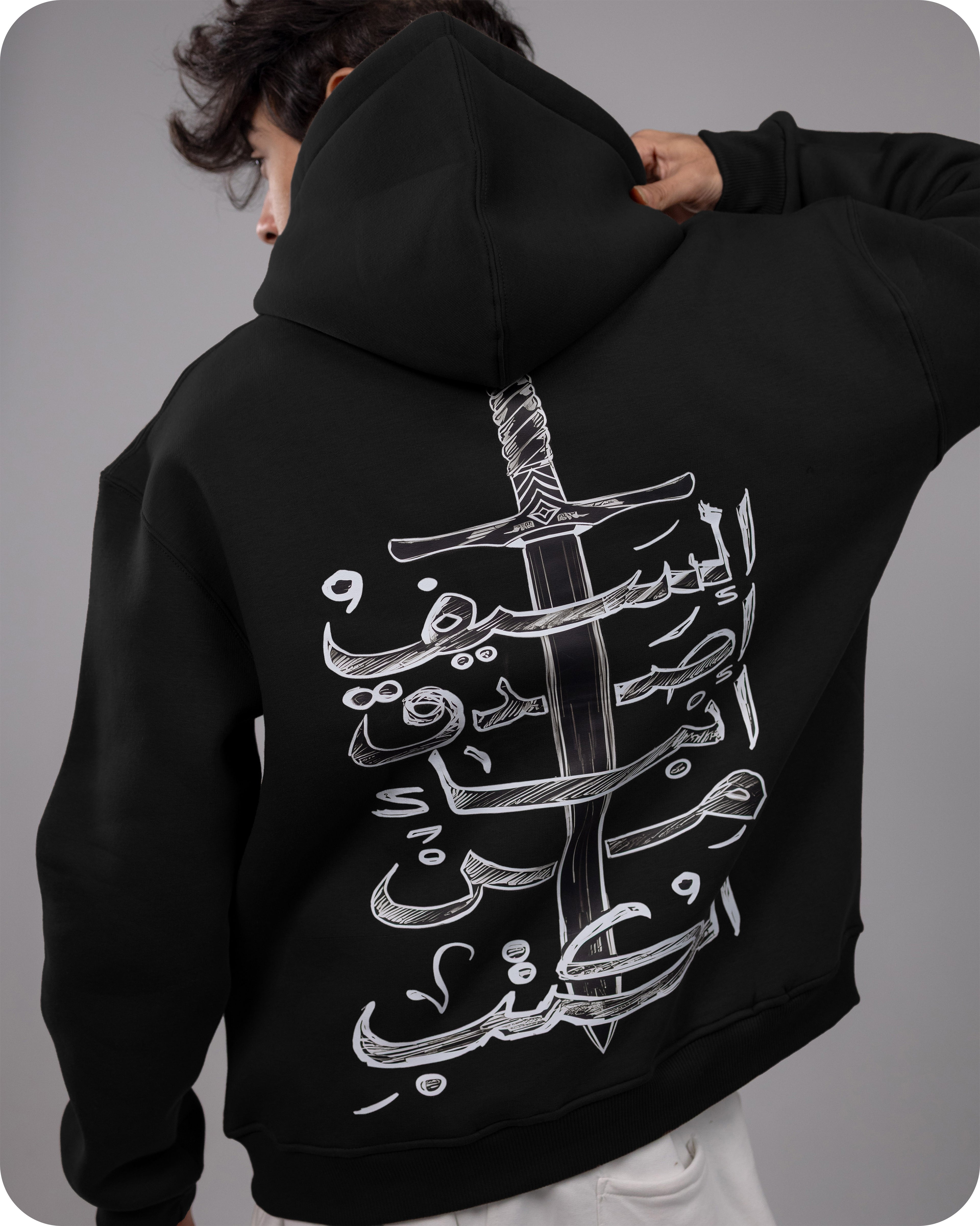 Person wearing a black hoodie with white Arabic calligraphy design on the back against a gray background