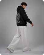 Person wearing a black hoodie with white text and white pants on a gray background