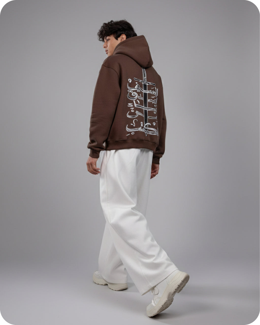 Person wearing a brown hoodie with Arabic text on the back, standing against a gray background
