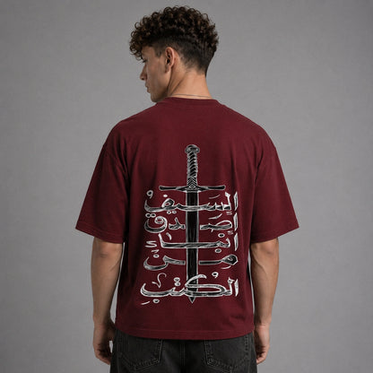 A man viewed from behind wearing a maroon t-shirt with a large graphic print of a sword and Arabic calligraphy on the back, paired with dark jeans against a gray studio background.