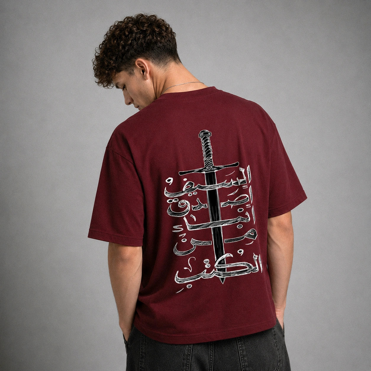 A man wearing a maroon t-shirt from the back, featuring a large graphic print of a sword and Arabic calligraphy. The background is a solid gray studio wall.