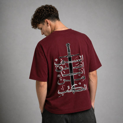 A man wearing a maroon t-shirt from the back, featuring a large graphic print of a sword and Arabic calligraphy. The background is a solid gray studio wall.