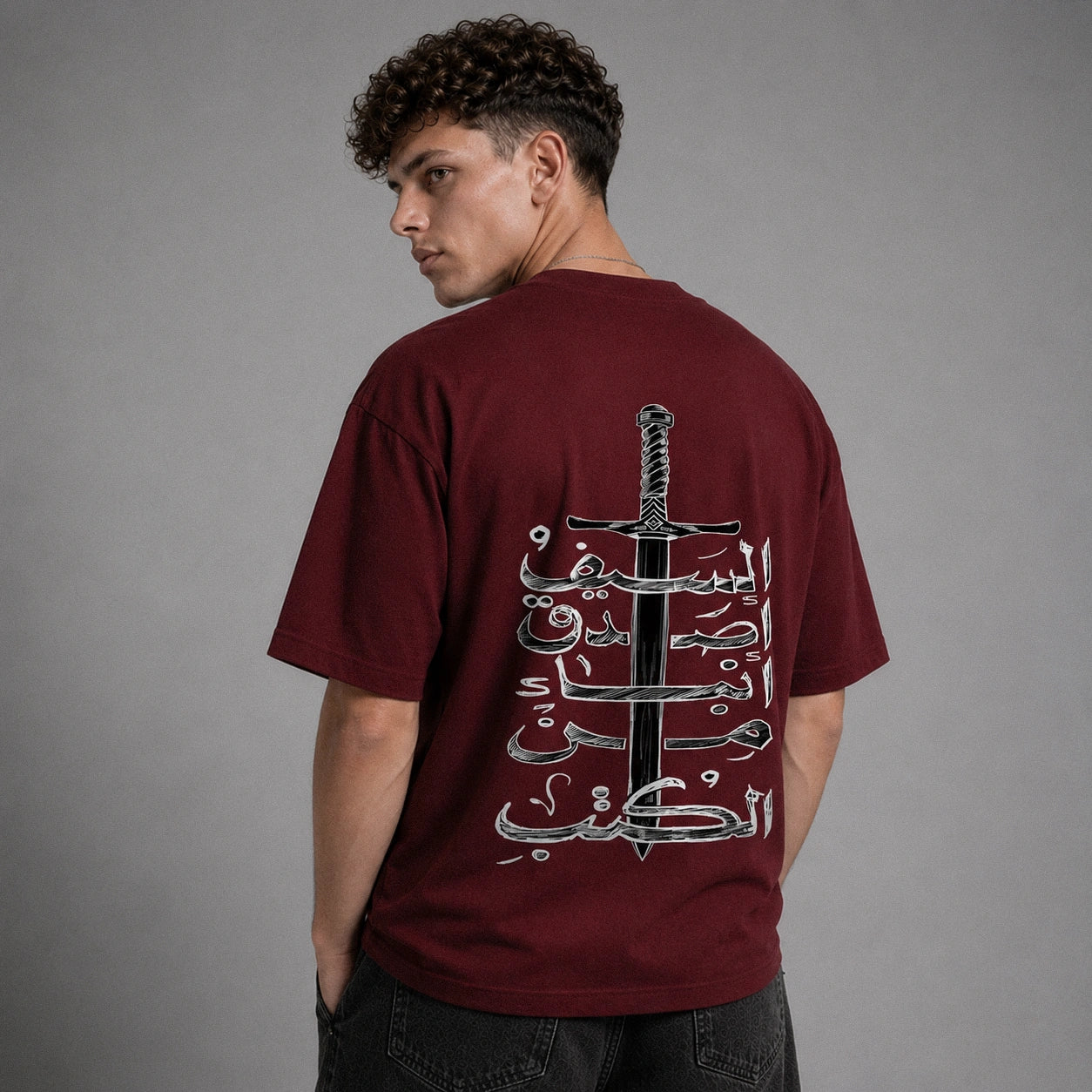 A man wearing a maroon t-shirt with a large graphic print on the back featuring a black sword and white Arabic calligraphy, photographed against a neutral gray background.