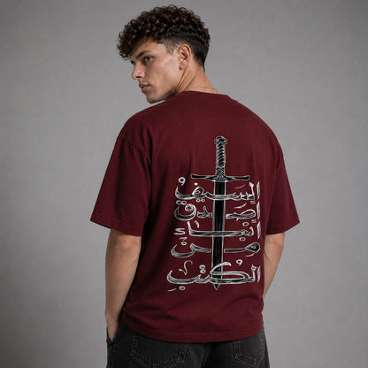 A man wearing a maroon t-shirt with a large graphic print on the back featuring a black sword and white Arabic calligraphy, photographed against a neutral gray background.