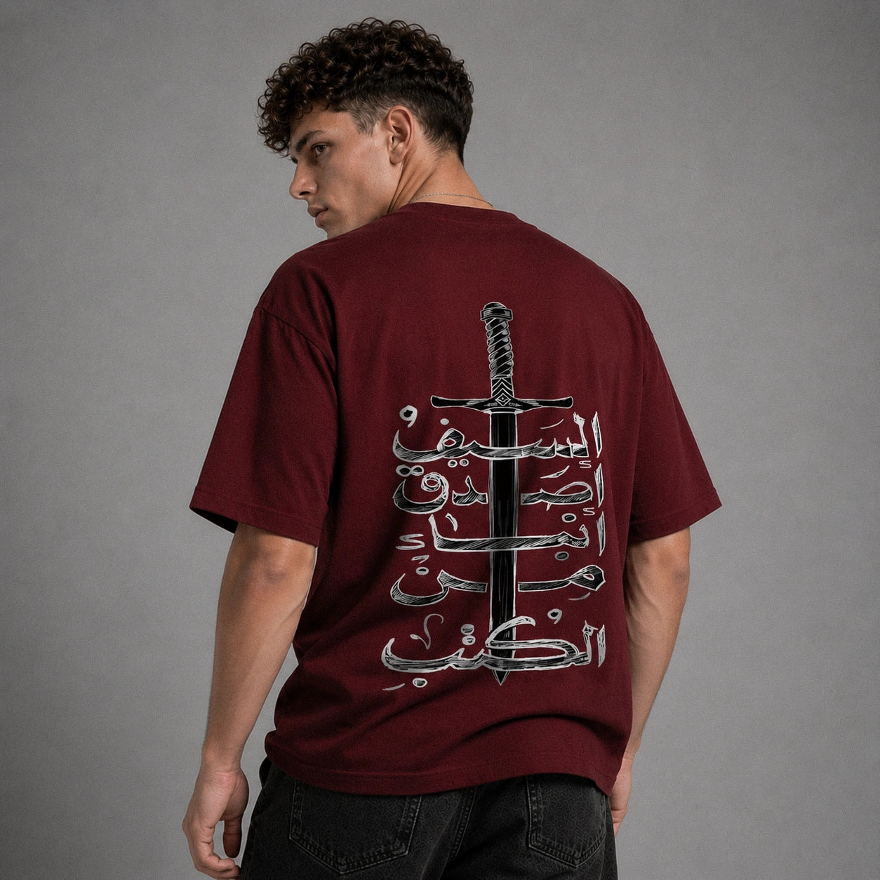 A man wearing a maroon t-shirt from the back, featuring a large graphic print of a sword and Arabic calligraphy in black and white. The background is a solid gray studio backdrop.
