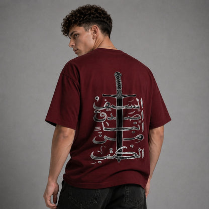 A man wearing a maroon t-shirt from the back, featuring a large graphic print of a sword and Arabic calligraphy in black and white. The background is a solid gray studio backdrop.
