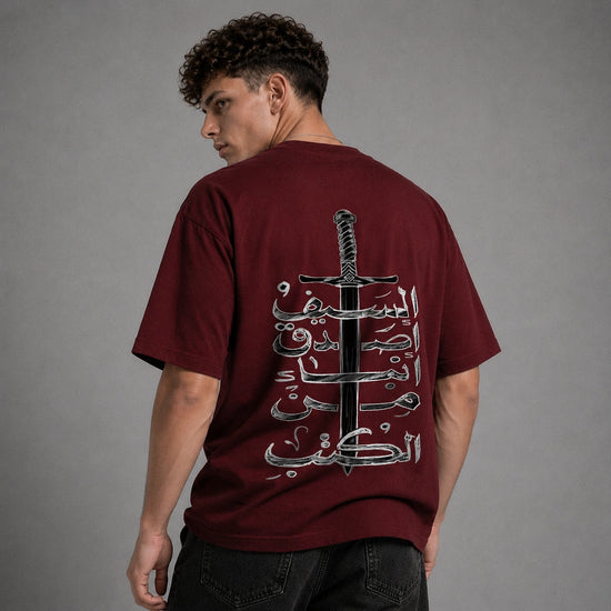 A man wearing a maroon t-shirt from the back, featuring a large graphic print of a sword and Arabic calligraphy in black and white. The background is a solid gray studio backdrop.