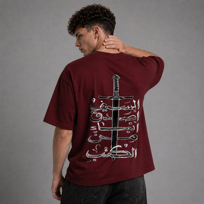 A man wearing a maroon t-shirt with a large graphic print of a sword and Arabic calligraphy on the back, shown from behind against a gray studio background.