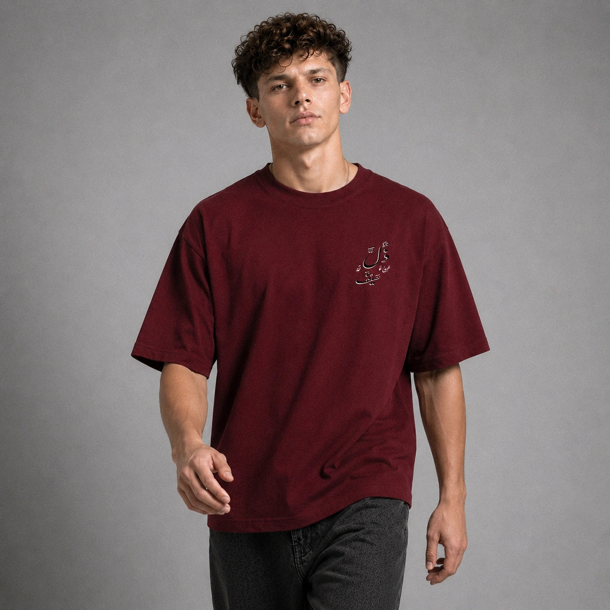 A man wearing a maroon crewneck t-shirt with a small embroidered logo on the left chest, paired with dark gray jeans, standing against a neutral gray studio background.