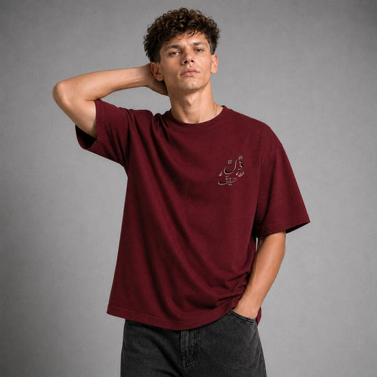A young man with curly brown hair wearing a maroon crewneck t-shirt with small white Arabic calligraphy on the left chest, paired with dark gray jeans, posing against a neutral gray studio background.