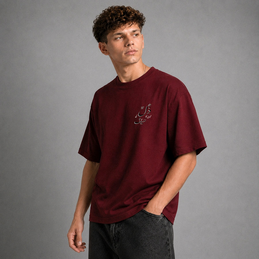 A man wearing an oversized maroon t-shirt with white Arabic calligraphy on the left chest, paired with dark gray jeans, standing against a solid gray studio background.