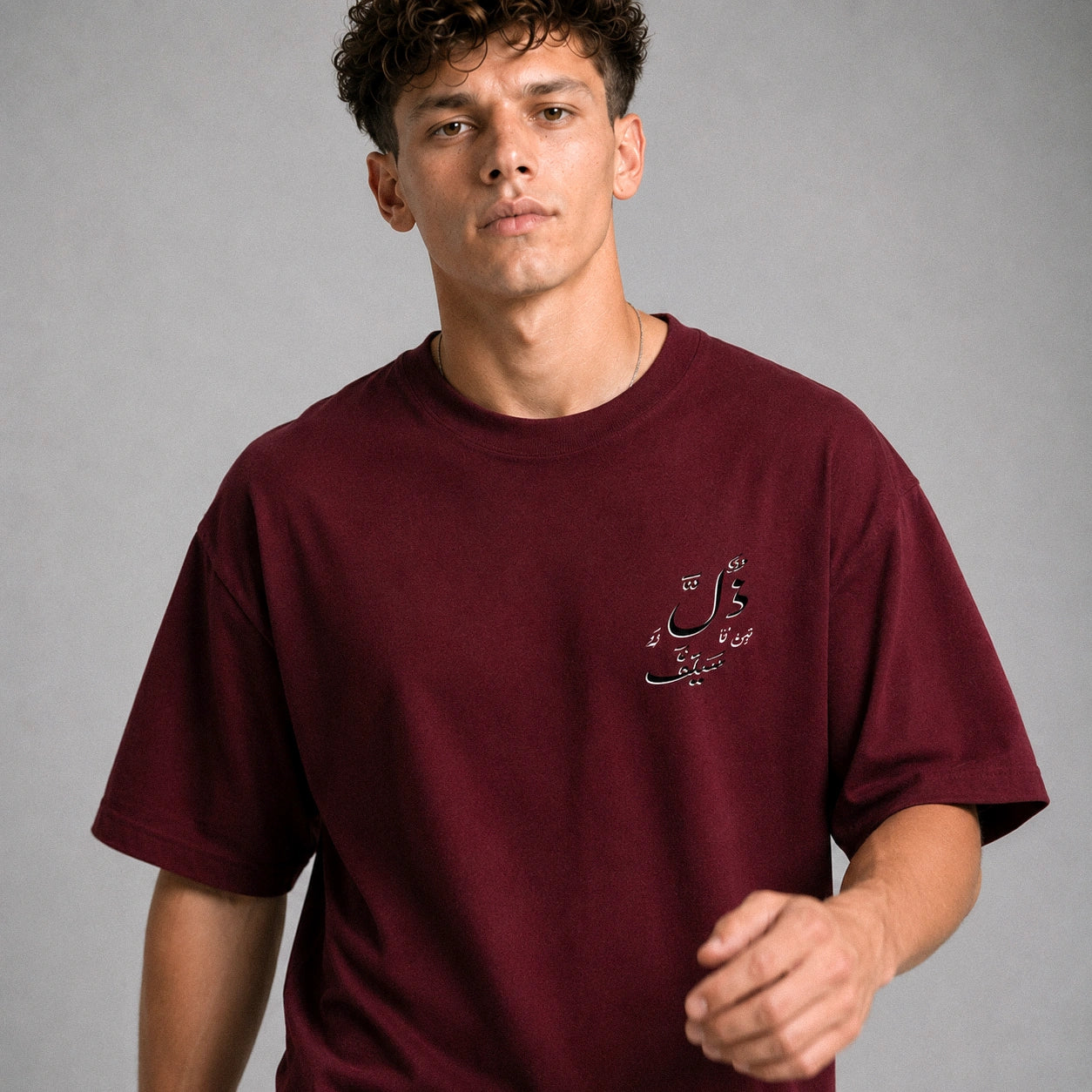 A man wearing a maroon crewneck t-shirt with a small black Arabic script graphic on the left chest, photographed against a neutral gray studio background.