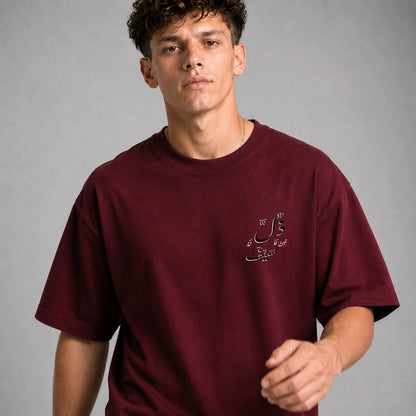A man wearing a maroon crewneck t-shirt with a small black Arabic script graphic on the left chest, photographed against a neutral gray studio background.
