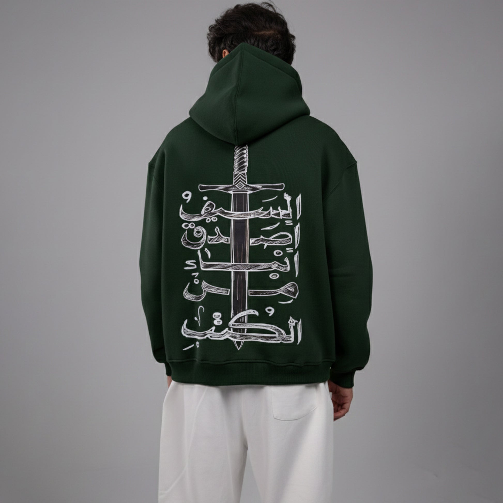 Person wearing a dark green hoodie with Arabic text design on the back against a gray background
