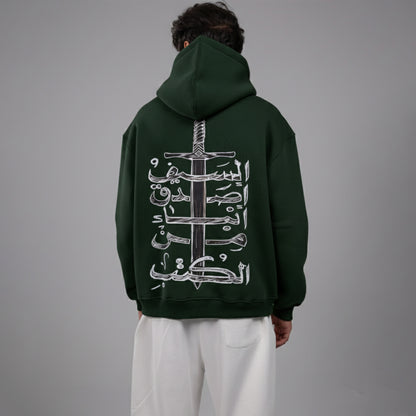 Person wearing a dark green hoodie with Arabic text design on the back against a gray background