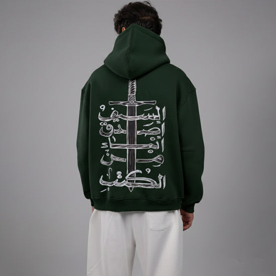 Person wearing a dark green hoodie with Arabic text design on the back against a gray background