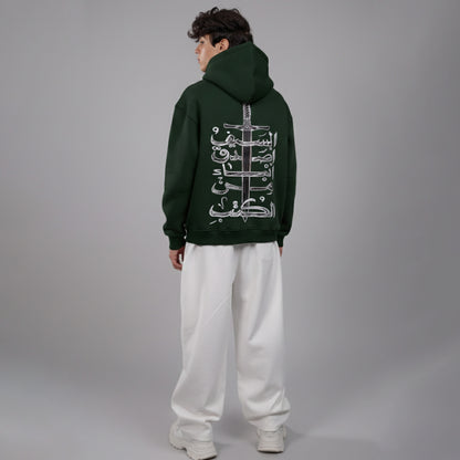 Person wearing a dark green hoodie with Arabic text on the back, standing against a gray background