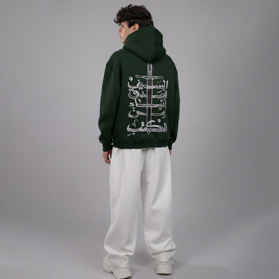 Person wearing a dark green hoodie with Arabic text on the back, standing against a gray background