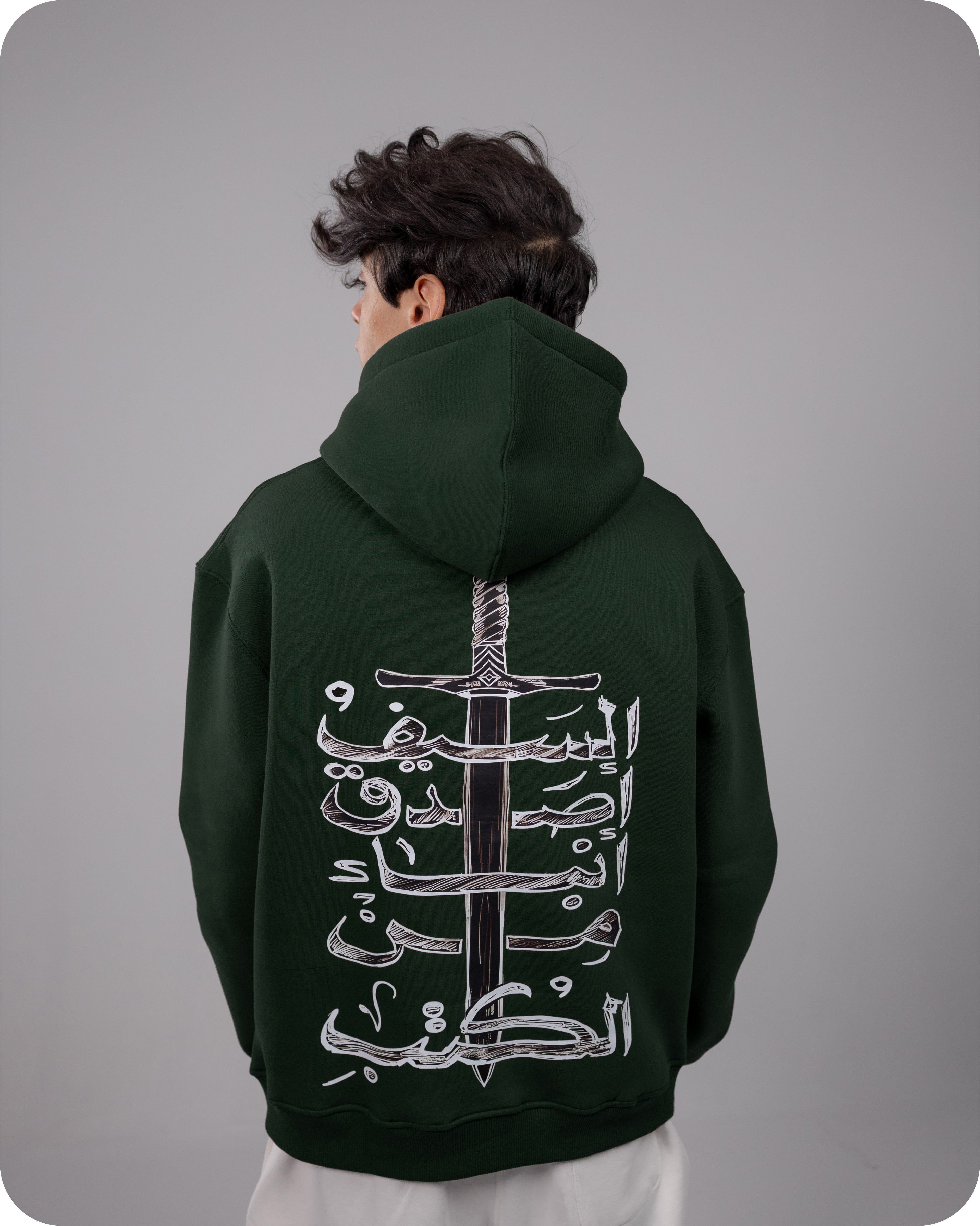 Person wearing a dark green hoodie with Arabic calligraphy design on the back against a gray background