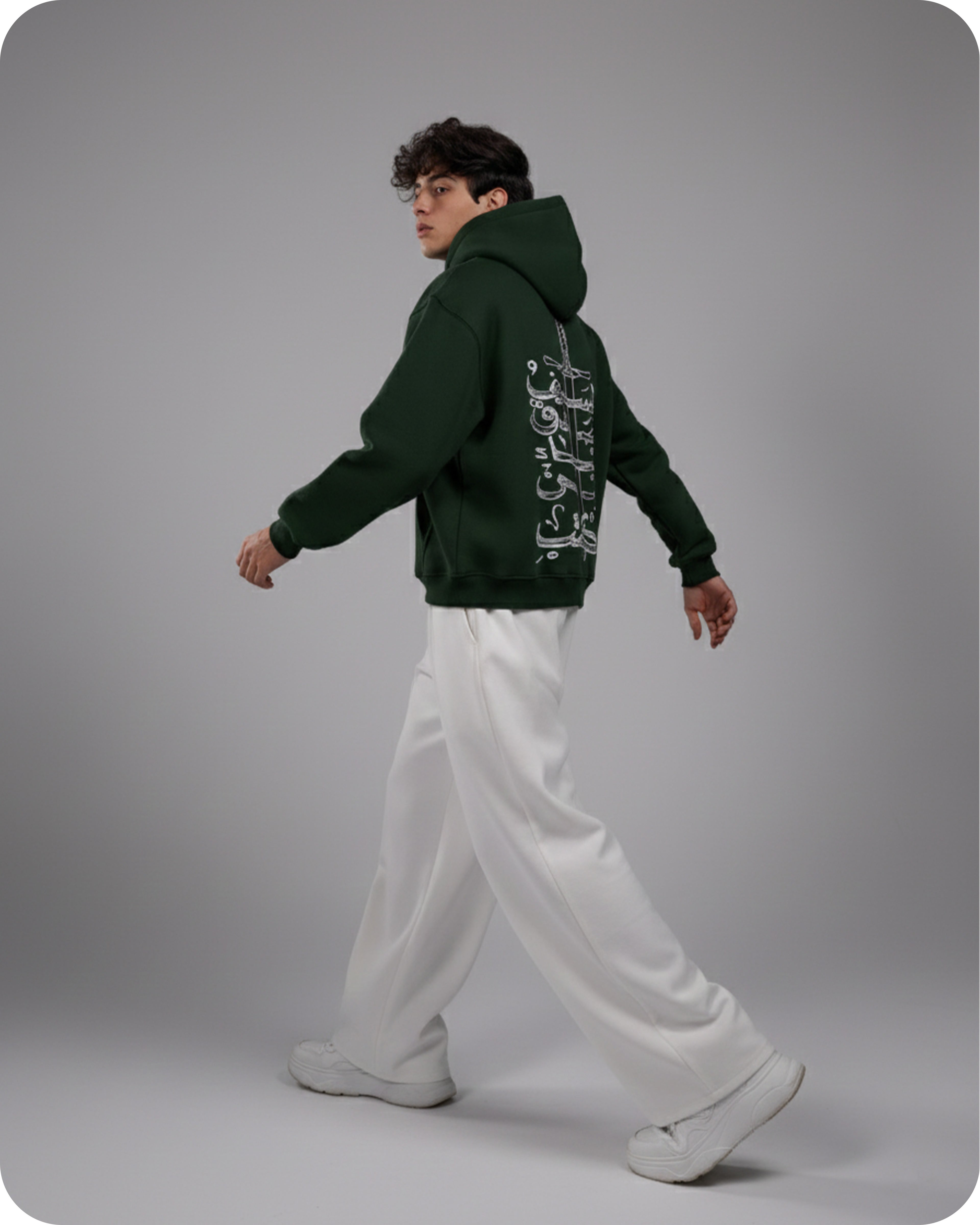 Person wearing a green hoodie with text on the back, white pants, and white shoes against a gray background