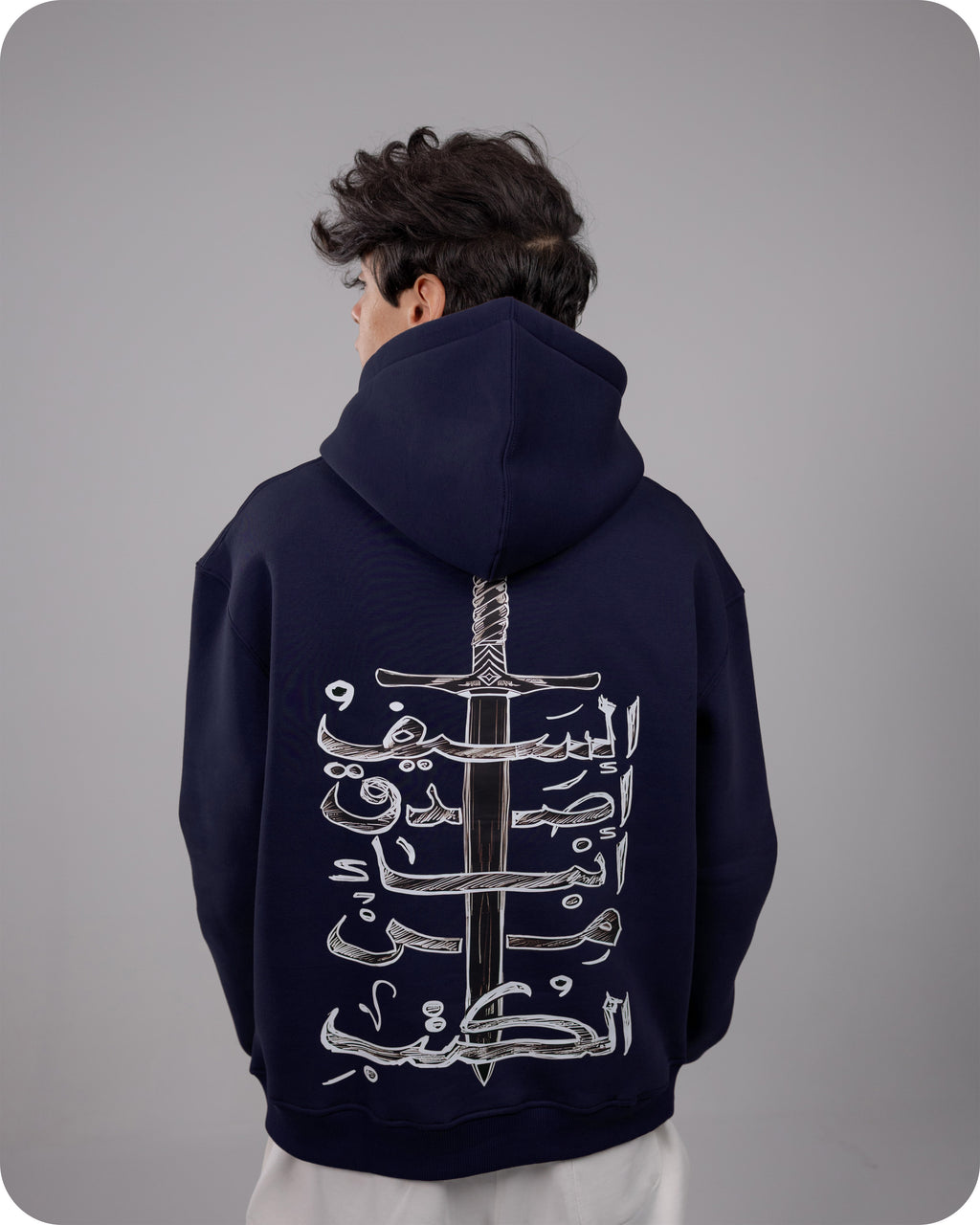 Person wearing a navy hoodie with white Arabic calligraphy design on the back against a gray background