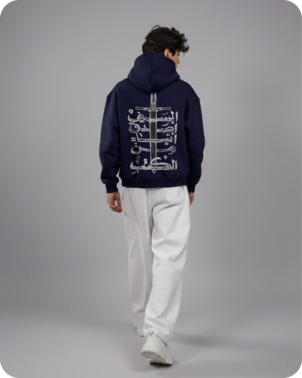 Person wearing a dark blue hoodie with white Arabic text on the back, standing against a gray background.
