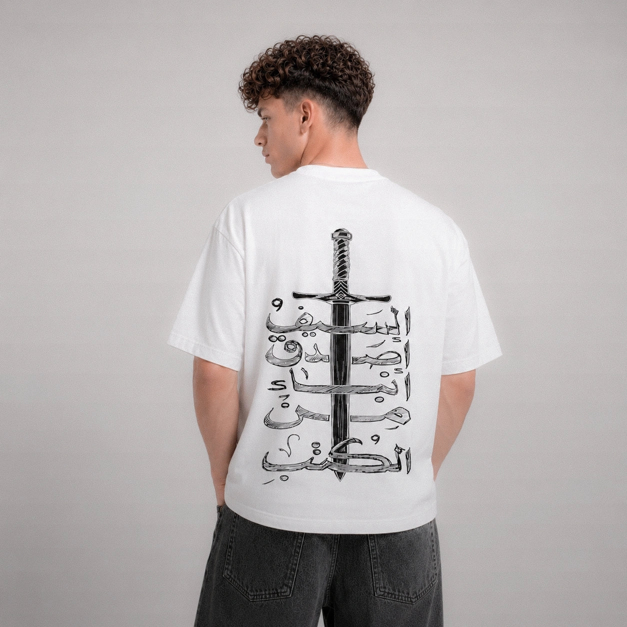 Person wearing a white t-shirt with a black sword and Arabic text design on the back, standing against a gray background.