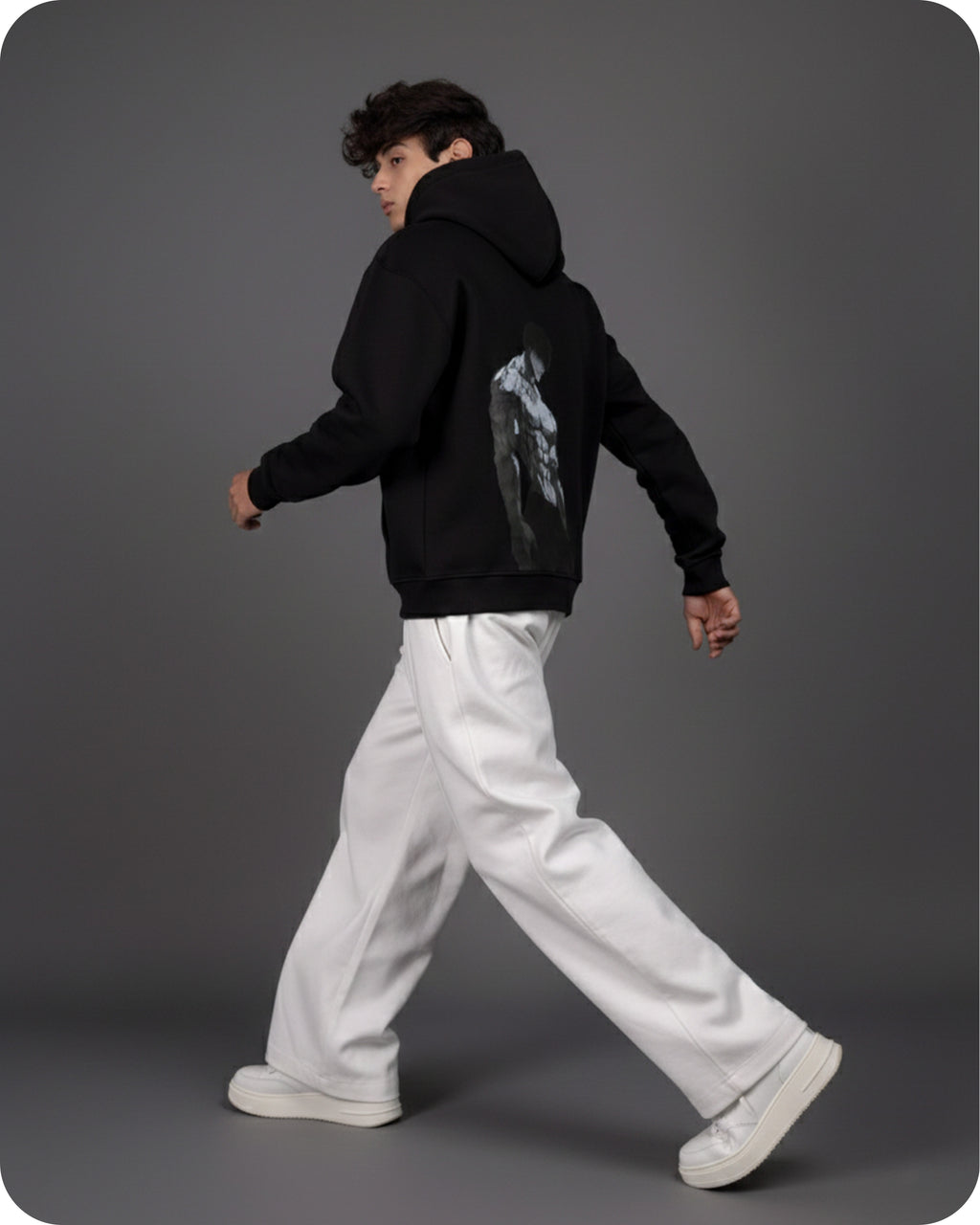 Person wearing a black hoodie with a graphic design on the back, white pants, and white shoes against a gray background