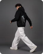Person wearing a black hoodie with a graphic design on the back, white pants, and white shoes against a gray background