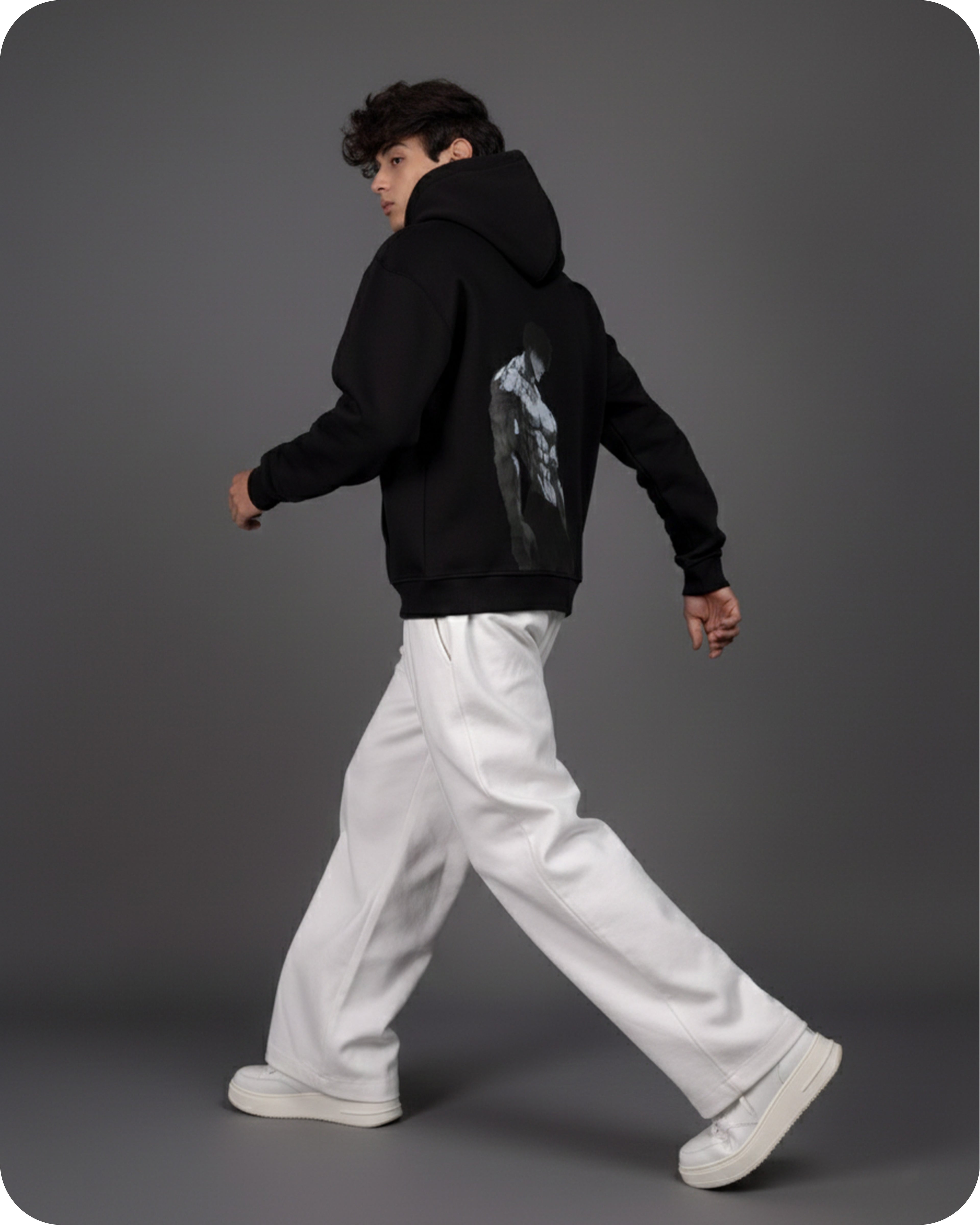 Person wearing a black hoodie with a graphic design on the back, white pants, and white shoes against a gray background