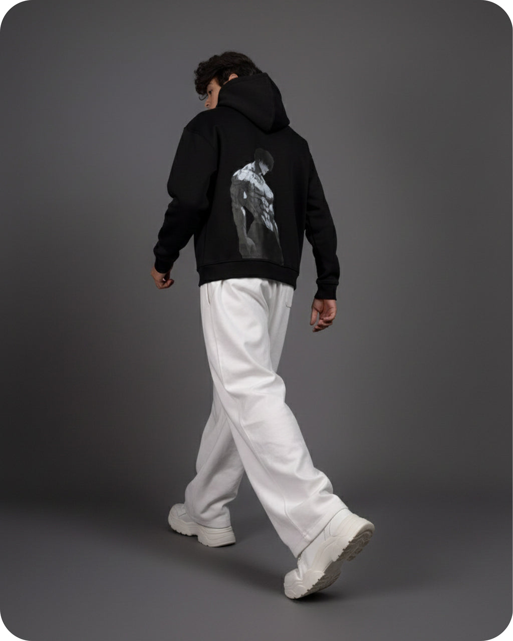 Person wearing a black hoodie with a graphic design on the back, white pants, and white shoes against a gray background