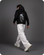 Person wearing a black hoodie with a graphic design on the back, white pants, and white shoes against a gray background