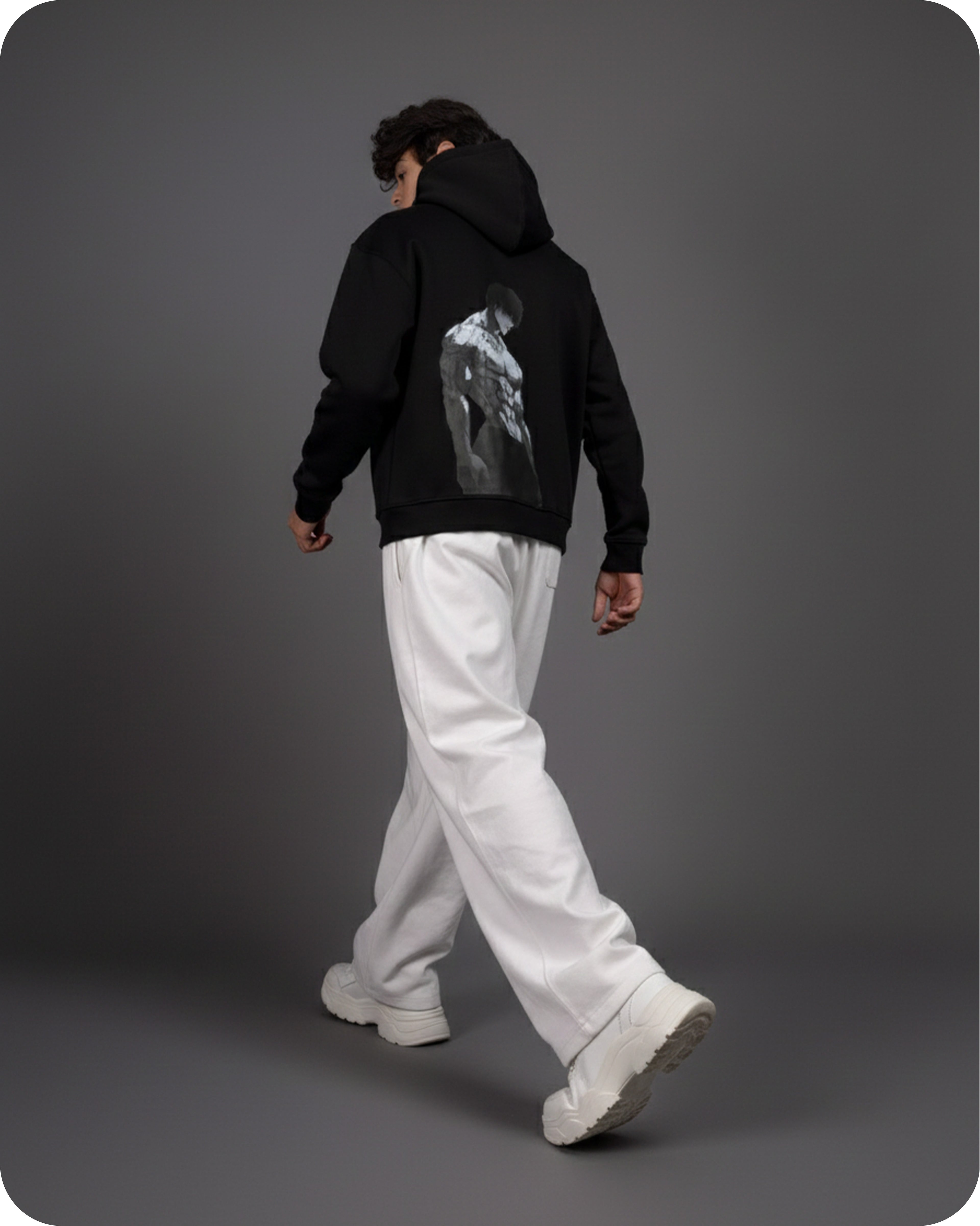 Person wearing a black hoodie with a graphic design on the back, white pants, and white shoes against a gray background