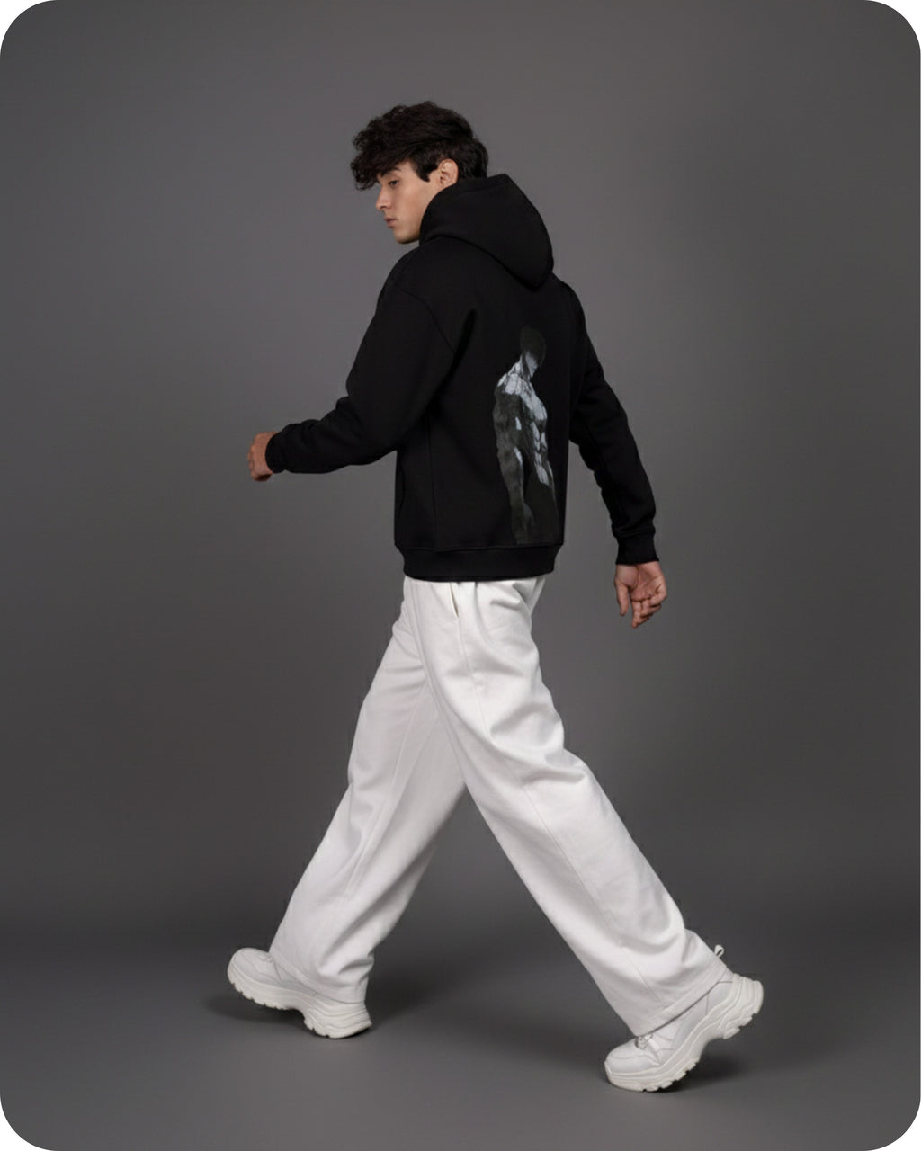 Person wearing a black hoodie with a graphic design, white pants, and white sneakers on a gray background
