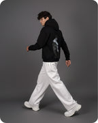 Person wearing a black hoodie with a graphic design, white pants, and white sneakers on a gray background